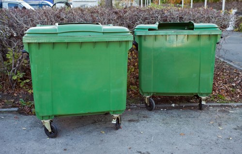 Two large green plastic wheelie bins with black lids are positioned side by side on a paved surface, likely a driveway or street in Lewisham. The larger bin on the left is slightly taller and has four visible wheels, one at each corner, facilitating movement. The smaller bin on the right also features a black lid and wheels but is shorter in height and narrower in width. Both bins are made of durable, textured plastic with a smooth finish, and are situated against a background of low, dense bushes with brownish foliage, indicating an autumn setting. The scene suggests rubbish collection or clearance activity typical in residential areas of Lewisham, possibly handled by local rubbish removal services like Lewisham House Clearance. The lighting appears natural, with soft shadows indicating daylight. The overall scene emphasizes the practical use of waste management containers in an urban UK residential context, supporting the environmental and housekeeping needs typical of the Lewisham postcode area.