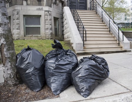 Three large black rubbish bags filled with waste are placed on the sidewalk near a tree in an urban area, adjacent to a building with a stone facade and small windows. The bags appear to be tied securely at the top and are situated close together, partially blocking the pavement. In the background, a set of concrete stairs with black metal handrails leads up to a higher level of the building, indicating a residential or commercial property in Lewisham. The surrounding environment includes a small patch of grass and a concrete driveway or pathway, typical of local streets where Lewisham House Clearance services might operate. The image captures a moment of rubbish collection or disposal, emphasizing the type of waste management activity that Lewisham House Clearance undertakes, particularly involving the removal of household or accumulated rubbish from local property clearances.