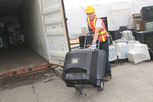 A worker in a yellow safety helmet and high-visibility vest is standing outdoors next to a partially open storage container or shipping unit, holding a large gray electronic device, possibly an old monitor or media equipment, by its handles. Behind the worker, there are various discarded electronic appliances and household items, including computer monitors and printers, placed on a paved surface. The background features a white covering or tent structure and a brick ledge, indicating a waste collection or clearance site, likely in Lewisham. The environment suggests a professional rubbish removal activity typical of Lewisham House Clearance services, with the worker preparing the electronics for disposal or recycling. The lighting appears natural, and the scene is clean yet cluttered with waste items awaiting collection.