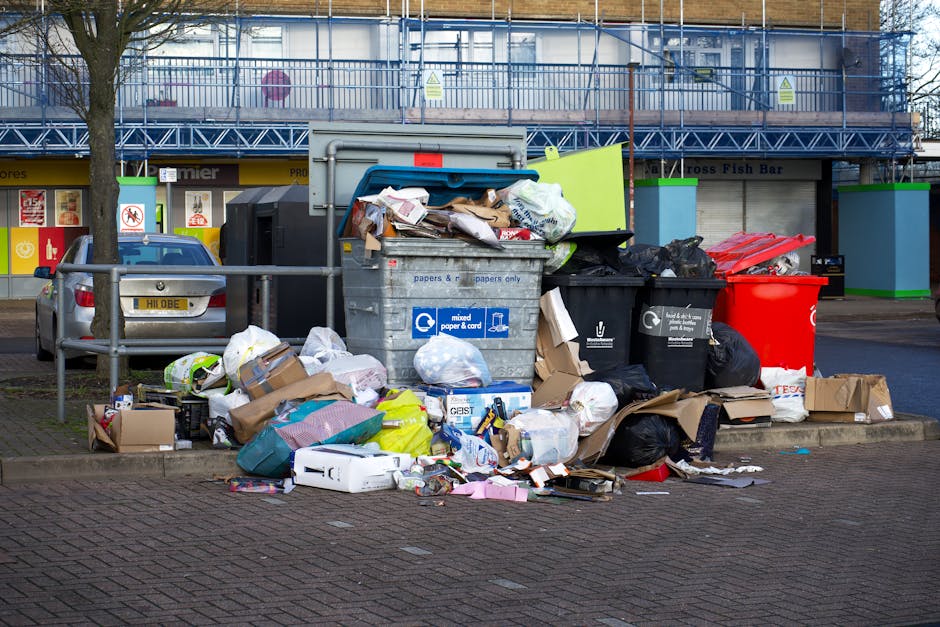 The image shows a pile of mixed waste and discarded materials situated outdoors on a paved surface, likely a driveway or yard area. The foreground features a heap of various items including large piec