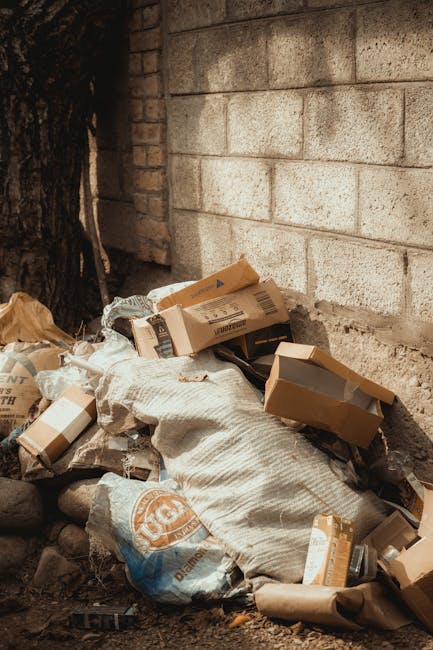 A black wheelie bin marked 'St. John's' is positioned on the pavement beside a street during nighttime. The bin is open and filled with various discarded items, including cardboard boxes, plastic, and
